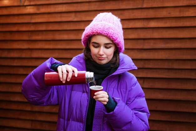 Une jeune femme heureuse boit du thé chaud tout en tenant un thermos un jour d'hiver boit pour se réchauffer dans...