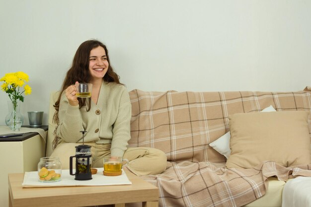 Jeune femme heureuse buvant une tisane dans une tasse en verre en détournant les yeux et souriant tout en étant assis sur un canapé