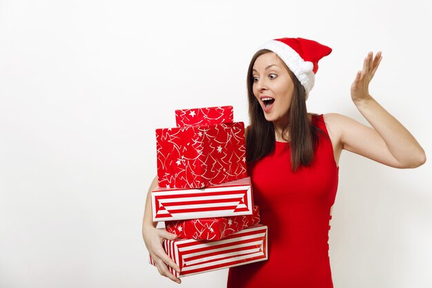Jeune femme heureuse caucasienne surprise avec un sourire charmant vêtue d'une robe rouge et d'un chapeau de Noël tenant des coffrets cadeaux sur fond blanc. Fille de Santa avec présent isolé. Concept de vacances de nouvel an 2018