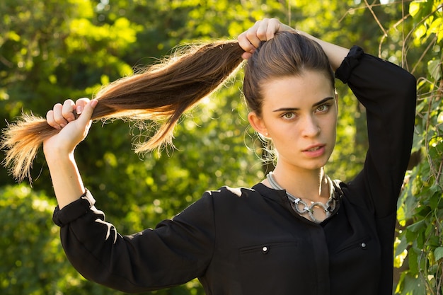 Jeune femme heureuse en chemisier noir touchant ses longs cheveux sur le fond des arbres verts