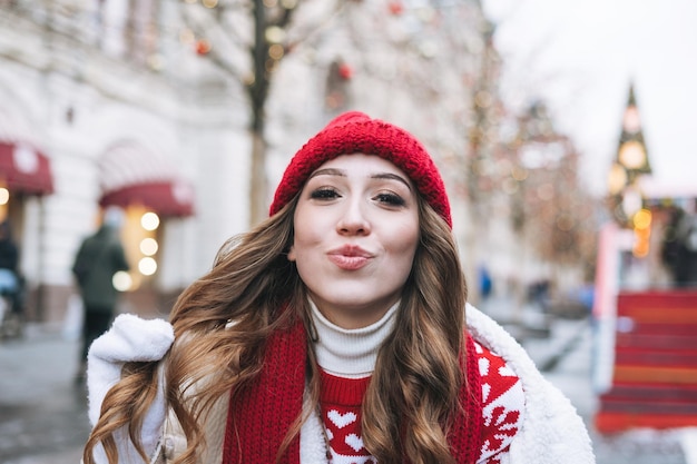 Photo jeune femme heureuse avec des cheveux bouclés sombres dans un chapeau tricoté rouge à la foire de noël dans la rue d'hiver