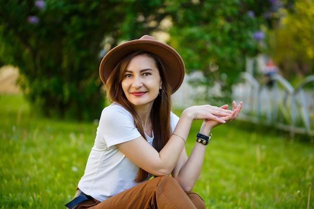 Jeune femme heureuse dans un chapeau est assise sur une pelouse verte dans un parc. Une fille d'apparence européenne avec un sourire sur son visage par une belle journée d'été ensoleillée