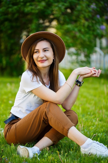 Jeune femme heureuse dans un chapeau est assise sur une pelouse verte dans un parc. Une fille d'apparence européenne avec un sourire sur son visage par une belle journée d'été ensoleillée