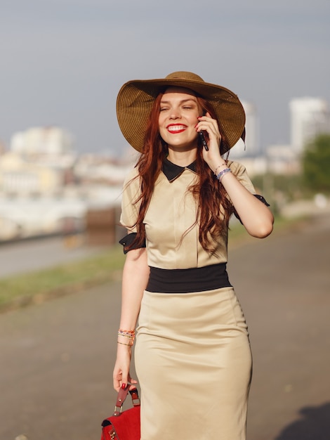 Une jeune femme heureuse dans un chapeau à larges bords marche dans le parc en été avec un sac et parle au téléphone