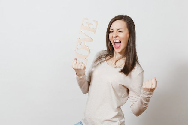 Jeune femme heureuse dans des vêtements légers et décontractés tenant un mot en bois amour, faisant le geste du gagnant sur fond blanc. Copiez l'espace pour la publicité. Concept de la Saint-Valentin ou de la Journée internationale de la femme.