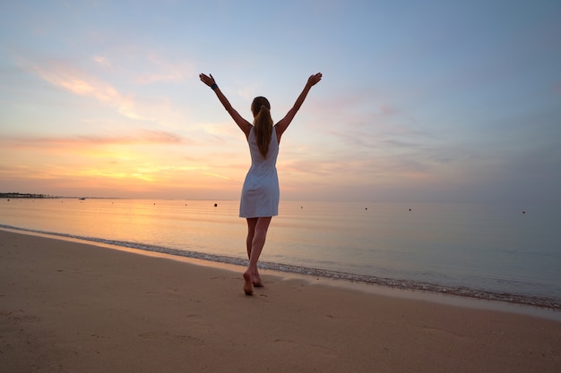 Jeune femme heureuse debout sur la plage de sable au bord de la mer profitant d'une chaude soirée tropicale.