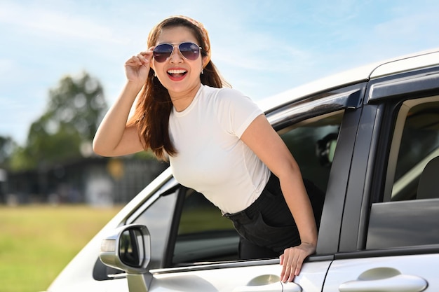 Une jeune femme heureuse et détendue avec des lunettes de soleil en route, en voyage, en vacances, penchée par la fenêtre de la voiture.