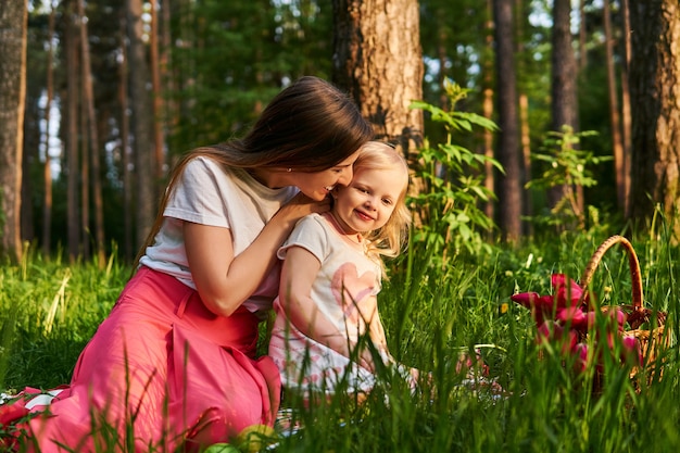 Une jeune femme heureuse embrasse son enfant lors d'un pique-nique dans la forêt à côté d'un panier de fleurs