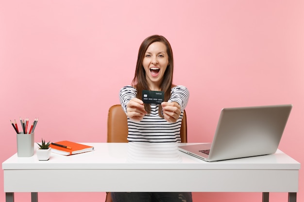 Une jeune femme heureuse et excitée tient une carte de crédit tout en étant assise, travaille au bureau au bureau blanc avec un ordinateur portable isolé sur fond rose pastel. Concept de carrière d'entreprise de réalisation. Copiez l'espace pour la publicité.