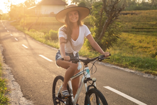Jeune femme heureuse fait du vélo par une route de campagne étroite