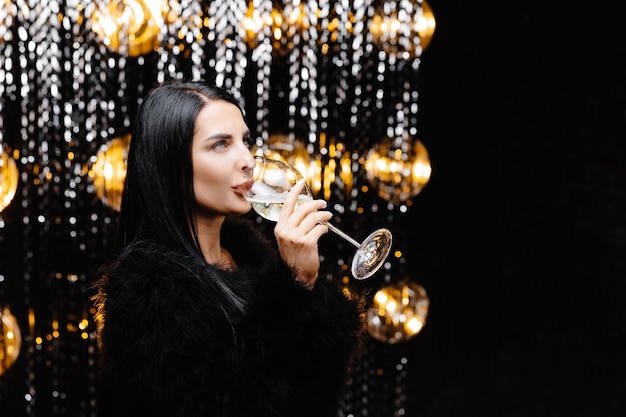 Jeune femme heureuse en manteau de fourrure de soirée branché regardant la caméra et souriant avec une coupe de champagne