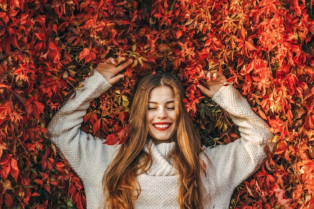 Jeune femme heureuse sur un mur de feuilles de lierre rouge. Elle sourit.