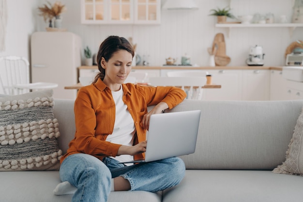 Une jeune femme heureuse avec un ordinateur portable est assise sur un canapé à la maison Travail à distance ou étude à domicile