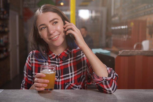 Jeune femme heureuse, parler au téléphone tout en buvant de la bière artisanale au pub