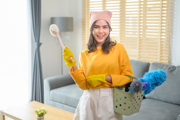 Jeune femme heureuse portant des gants jaunes et tenant un panier de produits de nettoyage dans le salon