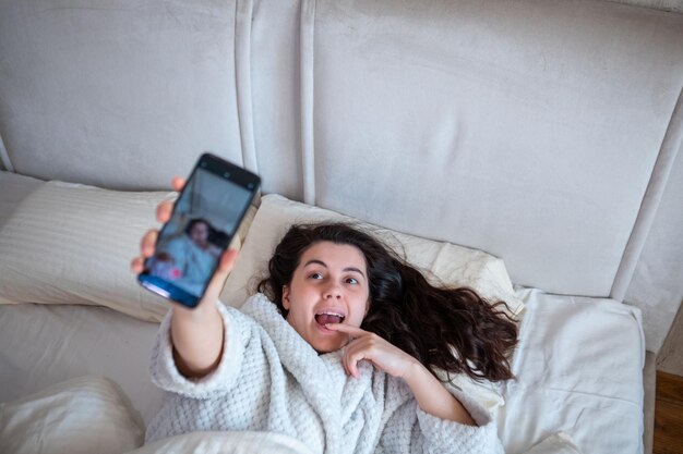 Jeune femme heureuse prenant une histoire de selfie allongée dans son lit