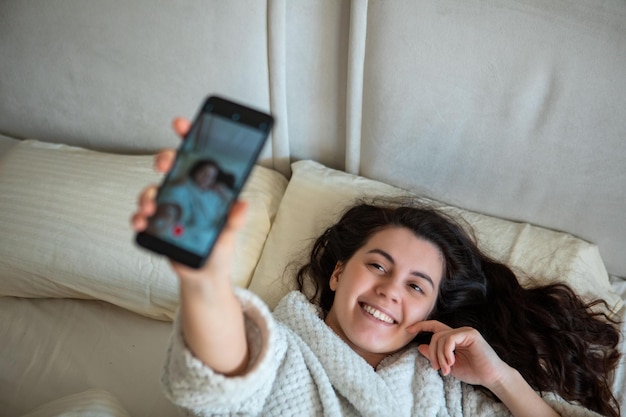 Jeune femme heureuse prenant une histoire de selfie allongée dans son lit