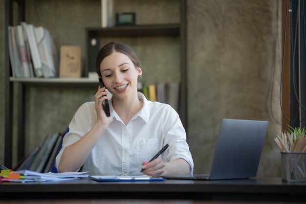 Une jeune femme heureuse qui parle sur son téléphone portable et sourit alors qu'elle est assise à son bureau.