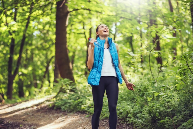 Jeune femme heureuse réussie après avoir couru un marathon avec une bouteille d'eau sur le fond du parc forestier naturel Sport