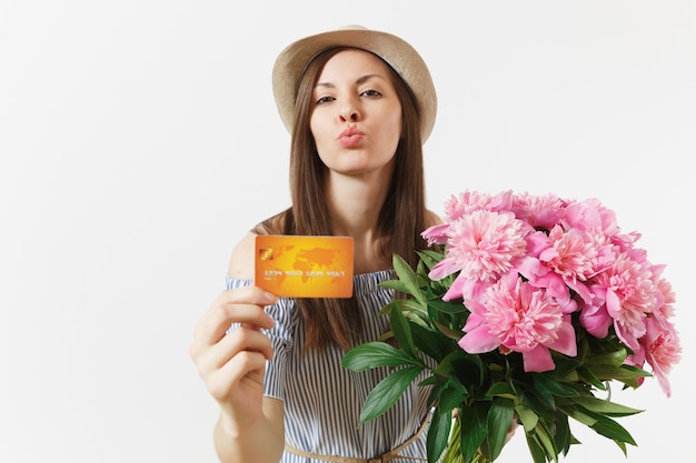 Jeune femme heureuse en robe bleue, chapeau tenant une carte bancaire de crédit, argent, bouquet de belles fleurs de pivoines roses isolées sur fond blanc. Affaires, livraison, concept d'achat en ligne. Espace de copie.
