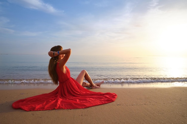 Jeune femme heureuse en robe rouge relaxante sur la plage de sable au bord de la mer profitant d'une chaude matinée tropicale.