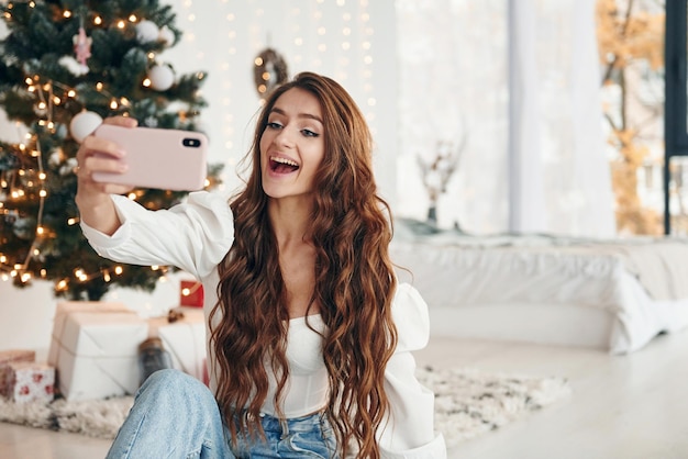 La jeune femme heureuse s'assied à l'intérieur près de l'arbre de Noël et célèbre la nouvelle année