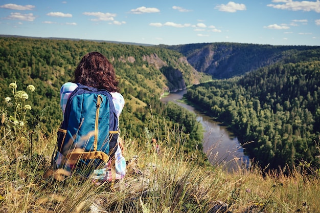 Jeune femme heureuse avec un sac à dos est assis sur une falaise et regarde la vallée ci-dessous