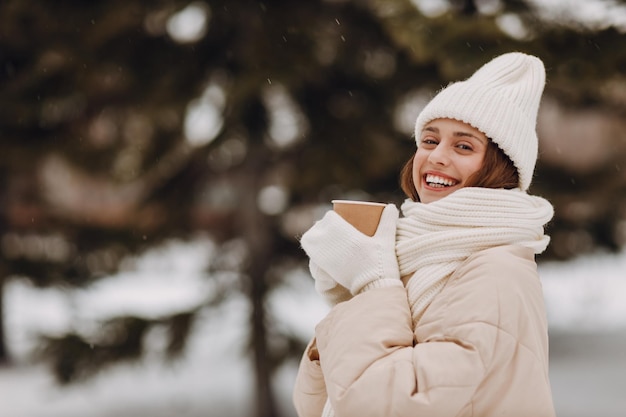 Jeune femme heureuse, souriante, portrait, manteau, foulard, chapeau et mites. Elle profite du temps hivernal dans un parc hivernal enneigé.