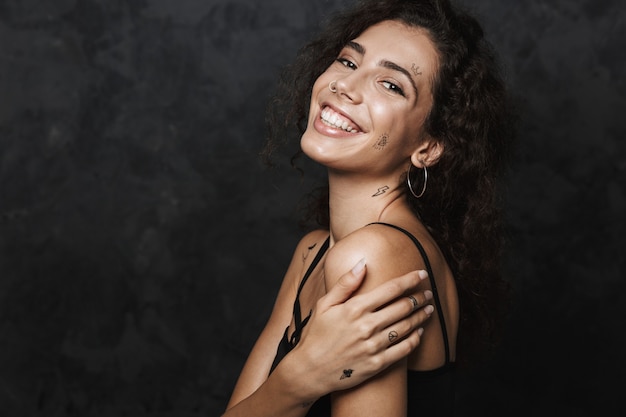 jeune femme heureuse avec un tatouage souriant et regardant isolé sur un mur noir