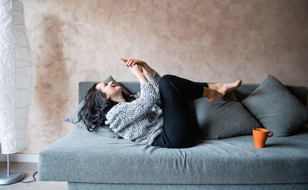 Une jeune femme heureuse utilisant un téléphone portable alors qu'elle est allongée sur le canapé à la maison