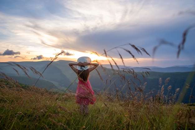 Jeune femme heureuse voyageur en robe rouge debout sur une colline herbeuse par une soirée venteuse dans les montagnes d'été, profitant de la vue sur la nature au coucher du soleil.