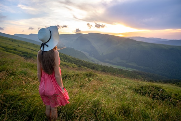 Jeune femme heureuse voyageur en robe rouge debout sur une colline herbeuse par une soirée venteuse dans les montagnes d'été, profitant de la vue sur la nature au coucher du soleil.