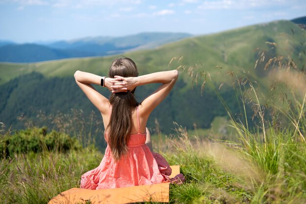Jeune femme heureuse voyageur en robe rouge reposant sur une colline herbeuse verte par une journée venteuse dans les montagnes d'été en profitant de la vue sur la nature