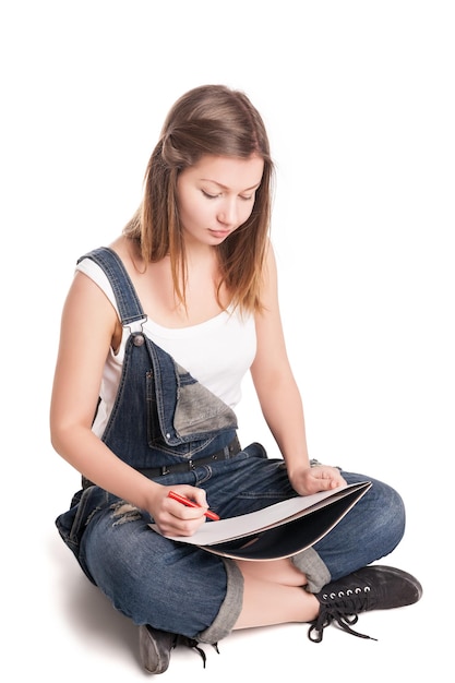 Jeune femme heureusement assise en tailleur sur le sol dessinant dans son bloc-notes sur fond blanc