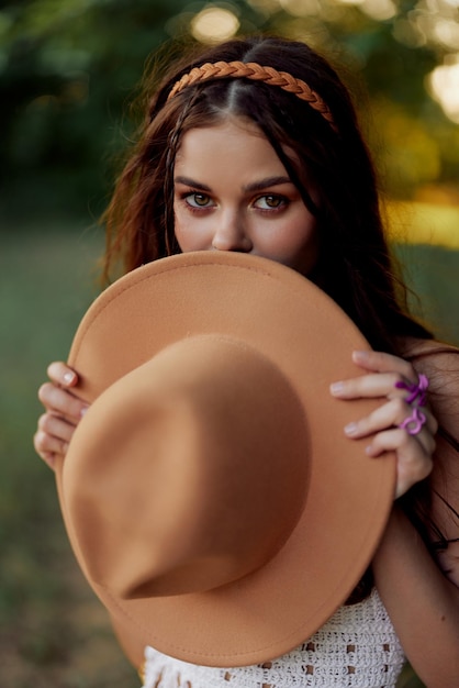 Jeune femme hippie tenant un chapeau et souriant sincèrement tout en regardant la caméra dans la nature à l'automne