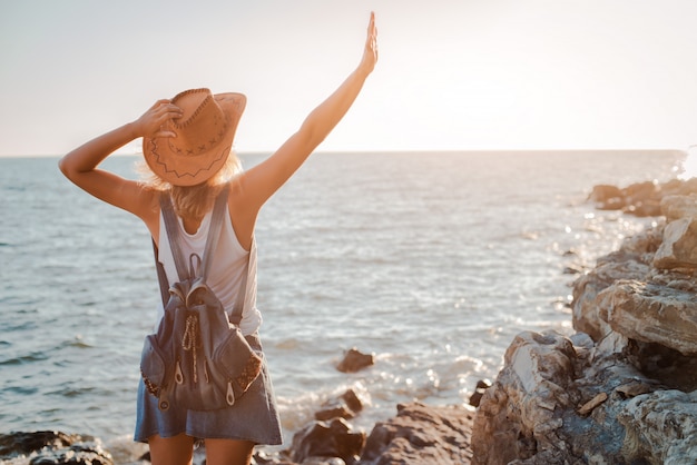 Une jeune femme hipster dans un chapeau et un rukzak avec ses mains, debout au sommet d'une falaise et regardant la mer au coucher du soleil.