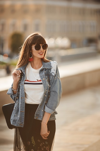 Jeune femme hipster dans une veste en jean et des lunettes de soleil rondes marchant dans la rue de la ville dans la journée d'été