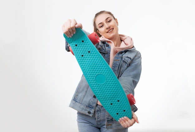 Jeune femme hipster gaie caucasienne dans des vêtements en denim avec penny board sur fond blanc