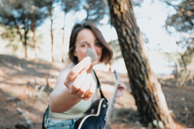 Jeune femme hipster jouant de la guitare en souriant à l'extérieur du parc forestier de la ville. En s'amusant à apprendre une nouvelle compétence, la musique joue un style saisonnier. Jeune fille aux cheveux courts. Espace de copie
