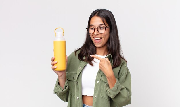 Jeune femme hispanique à l'air excitée et surprise pointant sur le côté et tenant un thermos à café