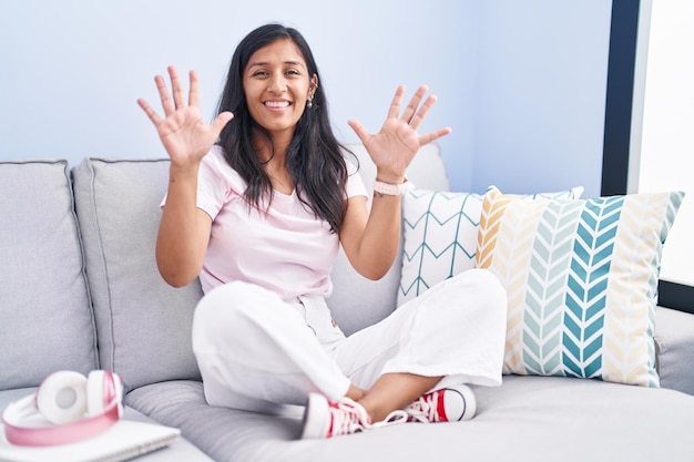 Jeune femme hispanique assise sur le canapé à la maison montrant et pointant vers le haut avec les doigts numéro dix tout en souriant confiant et heureux.