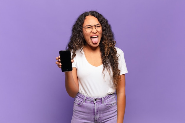 Jeune femme hispanique avec une attitude joyeuse, insouciante et rebelle, plaisantant et tirant la langue, s'amusant. concept de téléphone intelligent