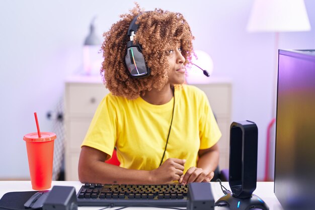 Jeune femme hispanique aux cheveux bouclés jouant à des jeux vidéo portant des écouteurs regardant sur le côté, pose de profil relaxante avec un visage naturel avec un sourire confiant.