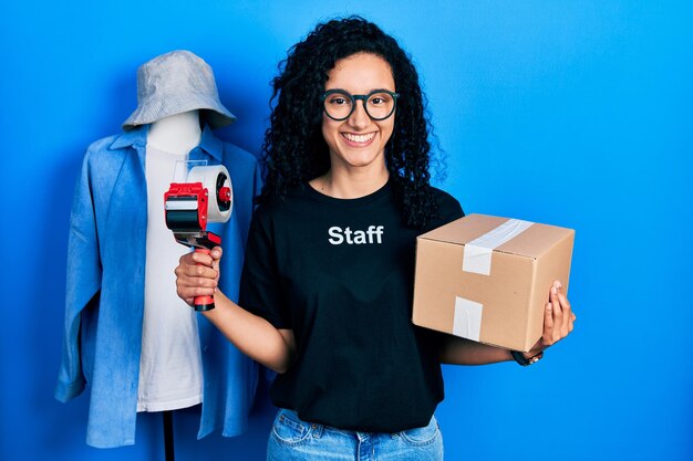 Jeune femme hispanique aux cheveux bouclés portant un t-shirt personnel tenant une boîte en carton souriant avec un sourire heureux et cool sur le visage. montrant les dents.