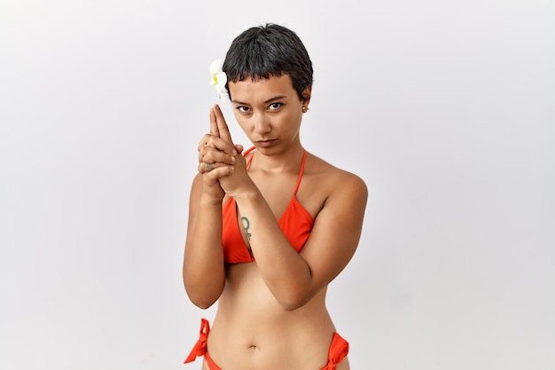 Jeune femme hispanique aux cheveux courts portant un bikini tenant un pistolet symbolique avec un geste de la main, jouant à tuer des armes de tir, un visage en colère