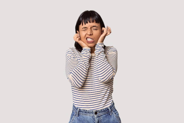 Une jeune femme hispanique aux cheveux noirs courts dans un studio couvrant les oreilles avec ses mains