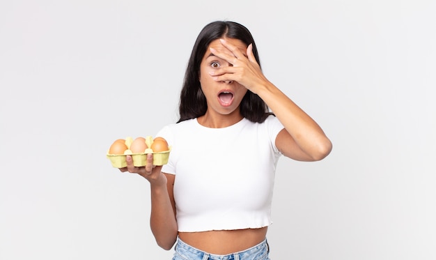 Jeune femme hispanique ayant l'air choquée, effrayée ou terrifiée, couvrant le visage avec la main et tenant une boîte à œufs