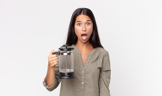 Jeune femme hispanique ayant l'air très choquée ou surprise et tenant une cafetière manuelle