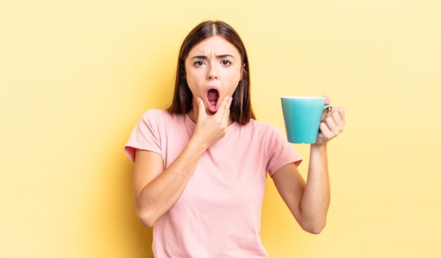 Jeune femme hispanique avec la bouche et les yeux grands ouverts et la main sur le concept de tasse de café de menton