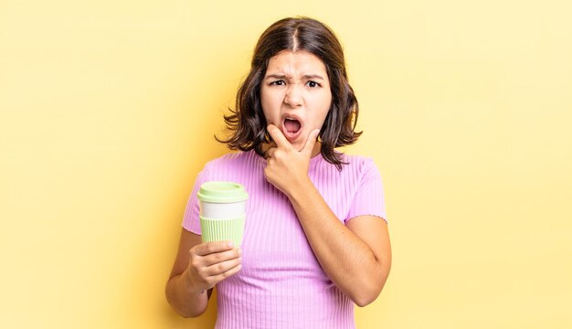 Jeune femme hispanique avec la bouche et les yeux grands ouverts et la main sur le menton emporter le concept de café